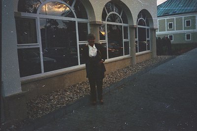 A man in a dark suit and patterned tie stands outdoors, reflected in large, arched windows. The building exhibits alpine arch...
