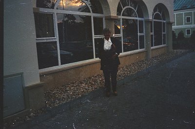 A woman in a dark suit and white shirt stands outside a building with a distinctive arched window facade. A gravel border sep...