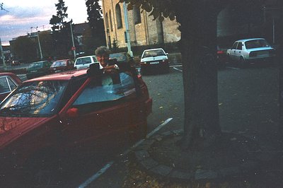 A man is framed within a maroon car, parked alongside a street with other vintage vehicles. The background reveals a building...