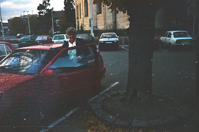 A vintage, grainy photograph depicts a man partially visible through the windshield of a red car, parked amongst other vehicl...
