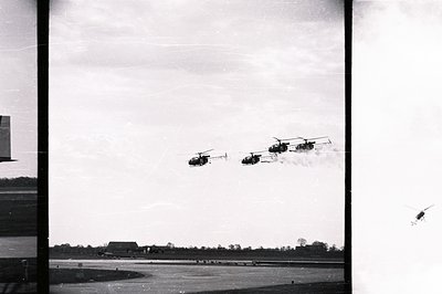 Four helicopters fly in formation across a rural landscape. Visible are treelines and a distant building, suggesting an airfi...