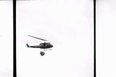 A black and white image shows a Bell UH-1 Iroquois ("Huey") helicopter in flight, carrying an external load. Possibly a resup...
