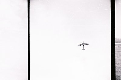 A high-contrast black and white image shows a lone paper airplane suspended mid-air against a hazy sky. The craft is simple i...