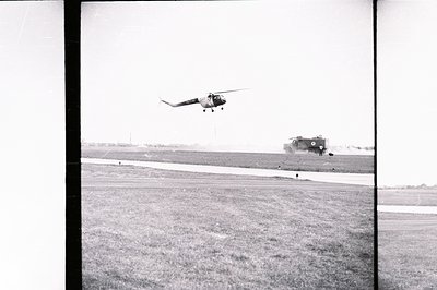 A Soviet-era helicopter hovers above a grassy airfield. Visible detail suggests a Mil Mi-4 model. Several figures are present...