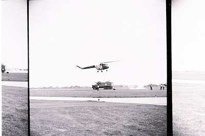 A Soviet-era Mi-4 helicopter hovers above an airfield, observed by personnel near a military vehicle. The scene suggests a tr...