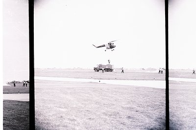 A military helicopter hovers above a grassy airfield, near a parked military transport vehicle. Several personnel are visible...