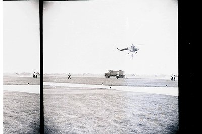 A military helicopter hovers over a grassy airfield with a rugged vehicle parked nearby. Several figures, possibly soldiers, ...