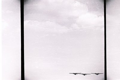 A trio of WWII-era aircraft, likely bombers, fly in formation against a cloudy sky. Visible grain and scratches indicate a po...