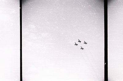 A vintage black and white photograph depicts four aircraft flying in formation across a bright sky. The image exhibits signif...