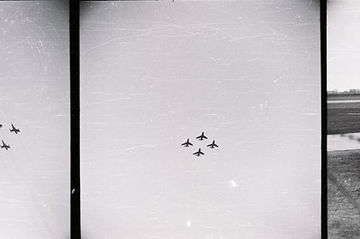 Four aircraft fly in formation across a clear sky, likely a military exercise or airshow. The landscape below shows fields an...