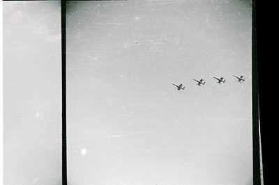 Four military aircraft fly in formation across a bright, overcast sky. The image exhibits significant photographic wear – vis...