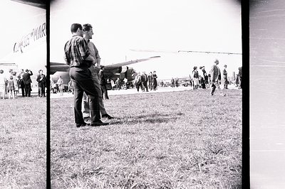 A scene at an airfield; two men in 1960s attire view an aircraft, likely an airshow. Several onlookers stand behind. Grass fi...