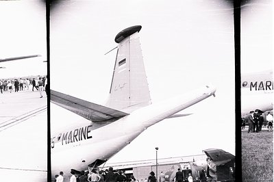 A black and white photo captures a large, military transport aircraft with “MARINE” marking clearly visible. Several people a...
