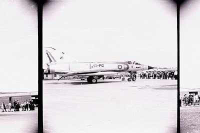 A Blackburn Buccaneer S.1 jet aircraft, marked "13-PG," taxis on a tarmac. A crowd of onlookers is visible in the background,...