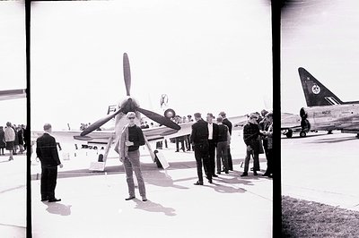 A group gathers around a Douglas DC-3 aircraft (registration NZ32) on an airfield. A man in a suit and trench coat stands pro...