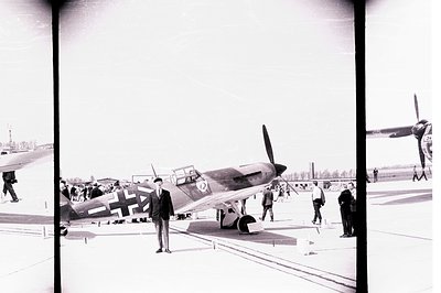 Vintage black and white photograph captures a Messerschmitt Bf 109 fighter plane, likely on display. A man in a suit and hat ...