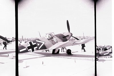 WWII-era Messerschmitt Bf 109 fighter plane sits on a tarmac with a few ground crew members present. The aircraft displays di...