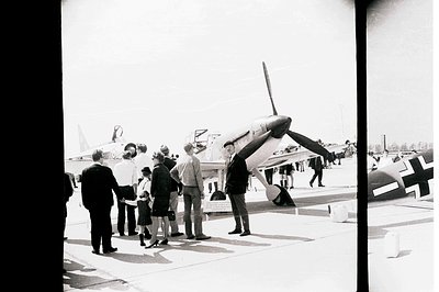 Group gathers around a restored Messerschmitt Bf 109 fighter plane at an airfield. Visible markings indicate a German militar...