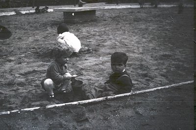 Two young boys in short trousers sit in a sandy play area, engaged in play. A third child is visible further away. The scene ...