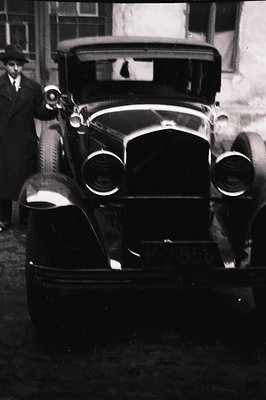 Classic 1930s automobile, likely a touring car, prominently features a visible license plate "P-7556." A figure in a suit sta...