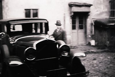 Vintage black and white image shows a man in a suit and hat standing next to a classic automobile, likely a 1930s model. Arch...