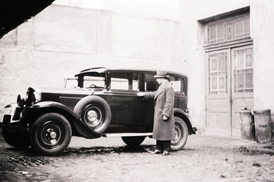 A gentleman in a long coat and hat stands beside a vintage 1930s automobile. The vehicle appears to be a pre-war sedan, likel...