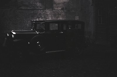 A black and white image depicts a vintage automobile, likely a 1930s model, parked in a shadowed courtyard. Visible details i...