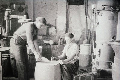 Two men work in what appears to be a photography darkroom. One, standing, adjusts equipment; the other sits, writing on a lar...