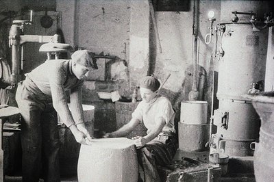 Two workers in a workshop, possibly leather or textile production. One is examining a stretched hide or fabric over a drum; t...