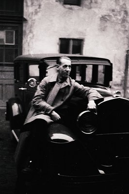 A man sits confidently on the front seat of a classic automobile, likely a 1920s or 1930s model. He wears a dark overcoat and...