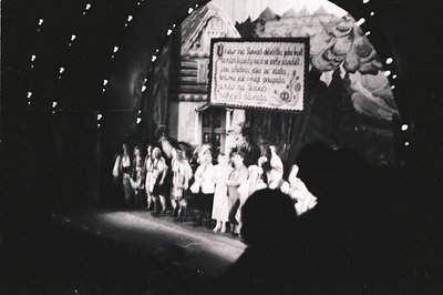 A group of people in traditional folk costumes performs in a staged, wooded area. The backdrop features a sign with text, pos...