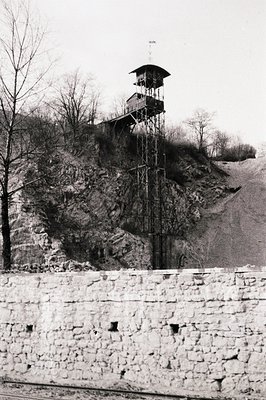 A stark, elevated watchtower rises above a stone wall, likely marking a border or restricted zone. The tower’s metal construc...