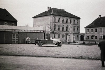 An elegant, open-top automobile stands before a three-story building with a central statue and symmetrical facade. A smaller ...