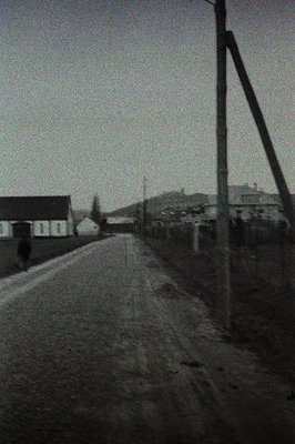 A stark, grainy black-and-white image captures a rural scene. A lone figure walks along a dirt road toward a distant village ...