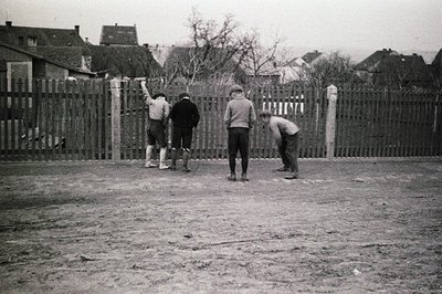 Three boys in caps and short trousers play in a muddy, enclosed yard, framed by a weathered wooden fence. Bare branches of a ...