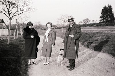 A formally dressed couple and a young woman stroll along a gravel road with a small terrier-like dog. The scene suggests an a...