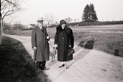 A formally-dressed couple strolls along a rural road with a small dog on a leash. The man wears a fedora and overcoat, using ...