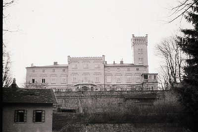 A stately manor house with a prominent tower rises from a terraced hillside, appearing slightly derelict. Classical architect...
