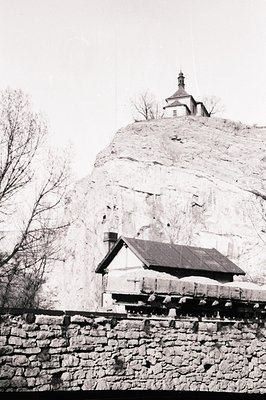 A stone building with a steep, gabled roof sits before a rocky, elevated terrain. A church with a distinctive spire crowns th...