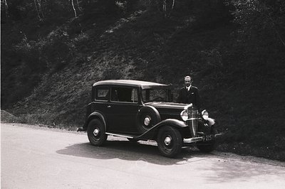 Elegant 1930s sedan, likely a Mercedes-Benz, stands on a rural road. A formally dressed man poses alongside. Forested hillsid...
