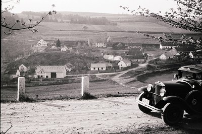 A vintage automobile is parked on a slightly raised, unpaved road overlooking a rural village. Farmhouses, barns, and a churc...