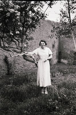 Elegant portrait of a woman in a polka-dot dress, posing near a stone wall and flowering tree. Likely 1930s fashion, judging ...