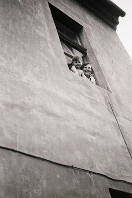 A black and white image captures two children peering out from a window set into a textured concrete wall. The children are s...
