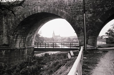 Stone arch bridge frames a view of a distant, ornate building with a spire, likely a church or castle. A small waterway flows...
