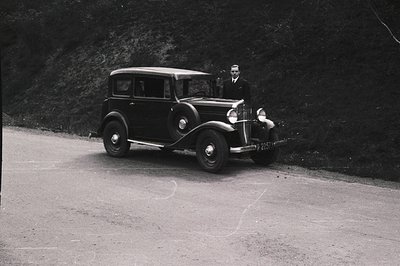 Classic black sedan, possibly a 1934 model, sits on a gravel road. A formally dressed man stands nearby. High contrast b&w ph...