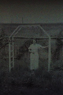 Black and white photograph featuring a man standing near a rustic wooden structure, possibly a trellis or small shelter. He w...