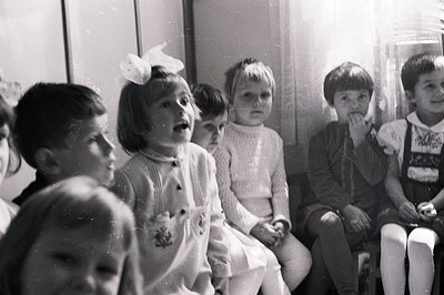 Group of young children, likely a class, seated indoors; attention focused on something beyond the frame. Clothing styles sug...