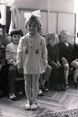A young girl in a dress & Mary Jane shoes stands prominently; other children seated in the background. A large bow adorns her...
