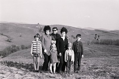 A family of six poses for a portrait on a grassy hillside, with a cable car visible in the background. The adults have charac...