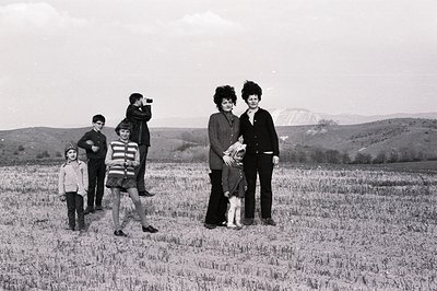 A family portrait, possibly Bulgaria, 1970s. A man holds a camera; three children and two women pose in a field. Women wear d...